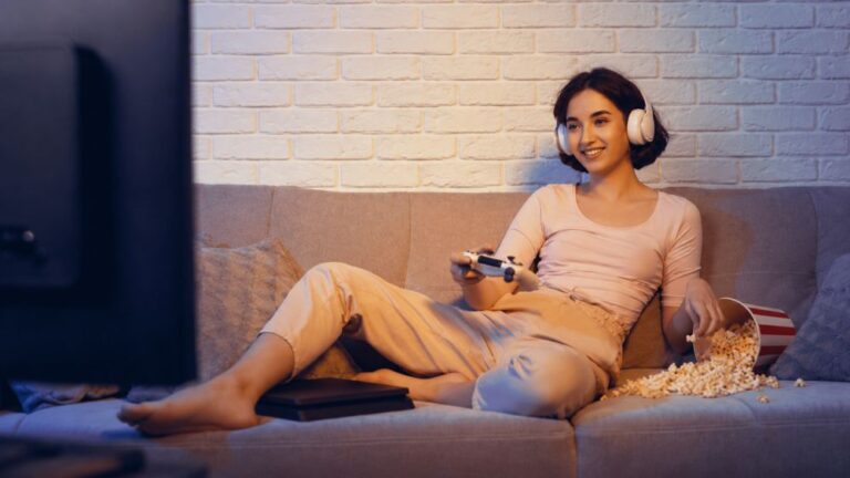 Devojka igra PlayStation konzolu na kauču sa slušalicama i kokicama